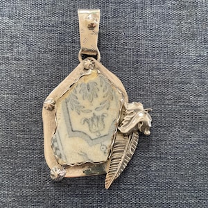May include: A silver pendant with a rectangular, light-colored stone center featuring a blue and white floral design. The pendant has a decorative silver feather and a floral design on the side.