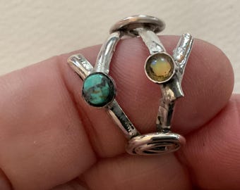 May include: Two silver rings with textured, branch-like bands. One ring features a round, blue-green turquoise stone, while the other ring is set with a round, iridescent opal stone. Both rings have a polished, metallic finish.
