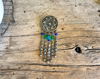 May include: A silver pendant with a circular design and blue and green beads hanging from it.