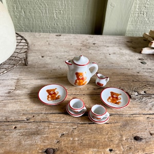 May include: A miniature tea set with a white teapot, two cups, two saucers, and two plates. The set features a brown bear design with red accents.