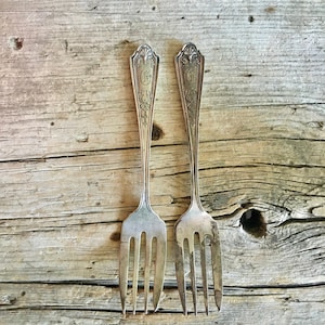 May include: Two silver forks with ornate handles and a decorative design. The forks are placed side by side on a weathered wooden surface.