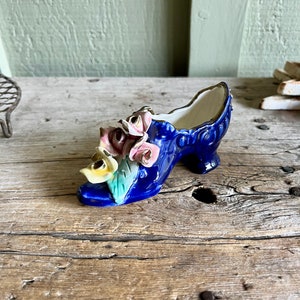 May include: A blue ceramic shoe with a floral design. The shoe is decorated with pink, yellow, and green flowers. The shoe is a trinket or decorative item.