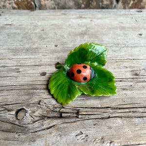 May include: A ceramic ladybug figurine with a red body and black spots rests on a glossy green leaf. The leaf has detailed veining and is slightly curved. The figurine is set against a rustic wooden surface.
