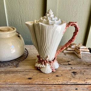 May include: A white ceramic teapot shaped like a seashell with a coral-colored handle. The teapot is decorated with raised details and sits on a wooden surface.
