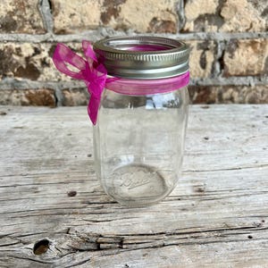 May include: A clear glass Ball jar with a silver lid and a pink ribbon tied around the neck. The jar is empty and sits on a weathered wooden surface. The background is a brick wall.