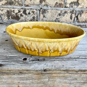 May include: An oval ceramic baking dish featuring a mustard yellow glaze with a white drip pattern cascading from the rim. The bowl has a smooth interior and a slightly ribbed exterior texture, showcasing a reactive glaze finish throughout the piece.