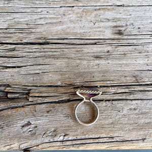 K&ouml;nnte beinhalten: Ein silberner Ring mit einem rechteckigen, lila Edelstein. Der Ring hat ein gedrehtes Seildesign um den Edelstein und ein einfaches, abgerundetes Band. Der Ring liegt auf einer verwitterten Holzoberfl&auml;che.
