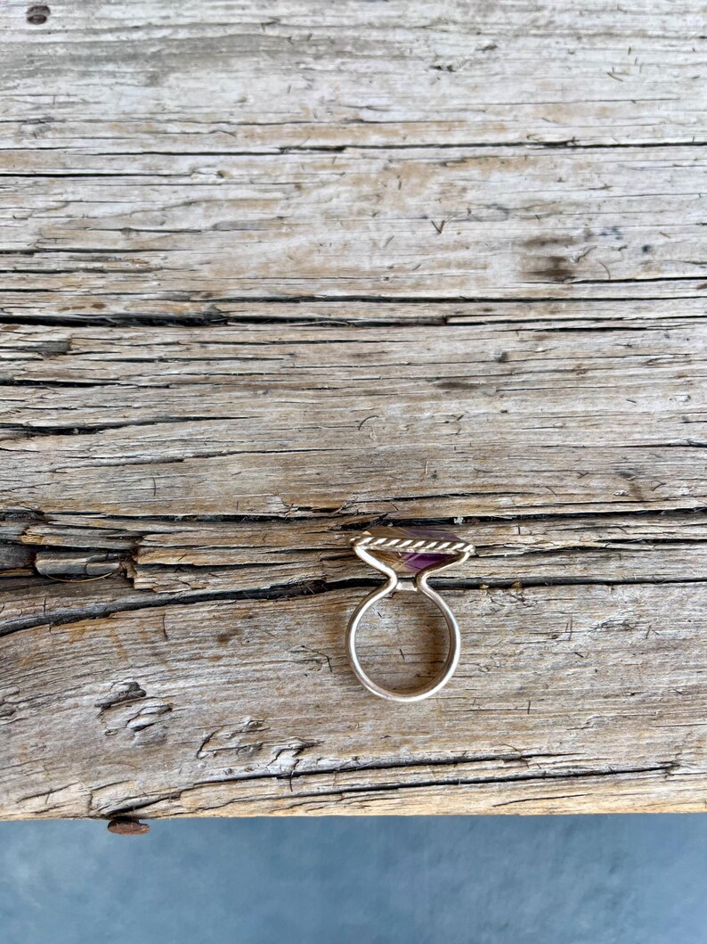K&ouml;nnte beinhalten: Ein silberner Ring mit einem rechteckigen, lila Edelstein. Der Ring hat ein gedrehtes Seildesign um den Edelstein und ein einfaches, abgerundetes Band. Der Ring liegt auf einer verwitterten Holzoberfl&auml;che.