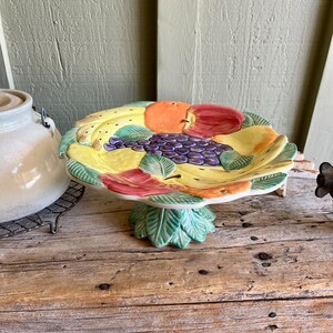 May include: A ceramic fruit bowl with a pedestal base. The bowl is decorated with a colorful design of apples, pears, bananas, and grapes.