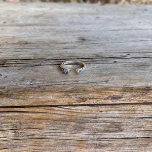 May include: A silver-toned open ring with a twisted band design. The ring features two light blue gemstones, one at each open end. The ring is set against a rustic wooden surface.
