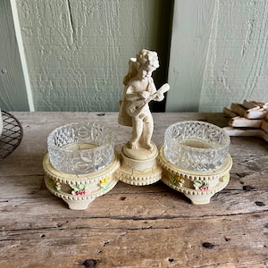 May include: A vintage double salt cellar with a cherub figurine playing a musical instrument. The salt cellars are made of clear glass with a cut design and sit on a white ceramic base with floral accents.
