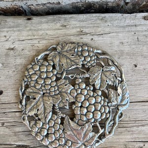 May include: A silver-colored, circular trivet with a grape and leaf design. The trivet features detailed clusters of grapes and large leaves, creating a decorative and functional kitchen accessory. The trivet is designed to protect surfaces from hot dishes.