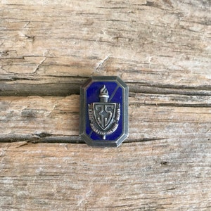 May include: A silver-toned pin with a blue enamel background. The pin features a shield with the text "35" and "PHILADELPHIA HIGH" inside. A torch is above the shield.