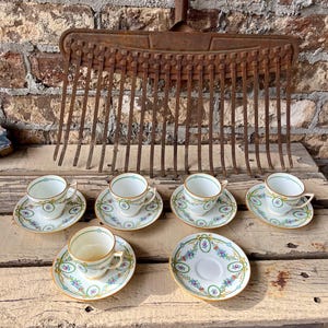 May include: Six white teacups and saucers with floral designs and gold trim are arranged on a wooden surface. An antique metal rake hangs in the background against a brick wall, creating a rustic aesthetic.