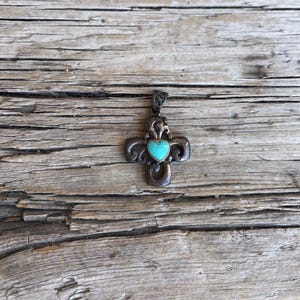 May include: A silver-toned cross pendant with an ornate design. The cross features a turquoise heart-shaped stone at its center. The pendant is displayed on a weathered wooden surface.