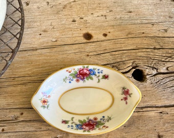 May include: A small, oval-shaped porcelain dish with a floral design. The dish has a gold rim and features a variety of pink, blue, and yellow flowers.