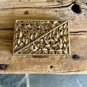 May include: A gold-colored metal box with a decorative design of leaves. The box has a hinged lid and a small latch.