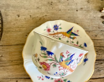 Op de afbeelding: Een wit theekopje en schotel met een gouden rand. Het theekopje en de schotel zijn versierd met een bloemenpatroon en een kleurrijk vogelontwerp.