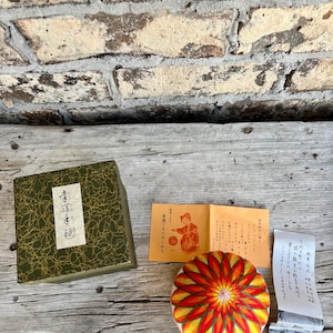 May include: An assortment of Japanese craft items, including a decorative box with gold accents, a colorful round Temari ball, and printed materials with Japanese text. The Temari ball features a vibrant red, orange, and yellow floral design.