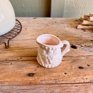 May include: A small white milk glass pitcher with a floral design. The pitcher has a pink interior and a handle.