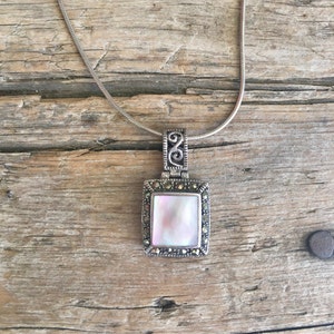 May include: A silver chain necklace with a square pendant featuring a white iridescent stone surrounded by a silver frame with black accents.