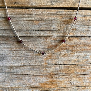 May include: A delicate silver necklace with small, round, deep red beads evenly spaced along the chain. The necklace is displayed against a rustic, weathered wooden background, highlighting its simple elegance.