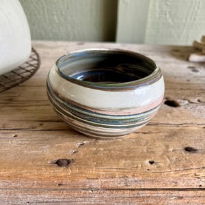 May include: A small, white ceramic bowl with a swirling pattern of brown, blue, and green stripes. The bowl is sitting on a wooden surface.