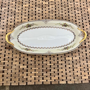 May include: An oval serving dish with gold handles and a floral design. The dish is white with a gold rim and a blue and green floral pattern.