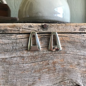 May include: A pair of silver earrings with a geometric, open-square design. The earrings are on a rustic wooden surface.