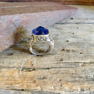 May include: A silver ring featuring a large, square-cut, deep blue gemstone. The ring has an ornate, filigree design around the gemstone and on the band. The ring is displayed on a weathered wooden surface.