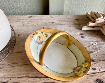 May include: A small, oval-shaped porcelain dish with a gold rim and floral design. The dish has a gold handle and is ideal for serving small snacks or treats.