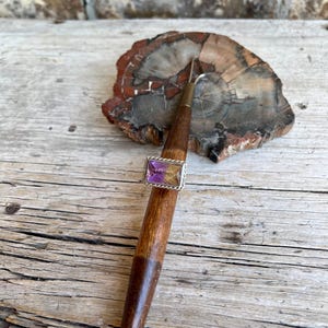 K&ouml;nnte beinhalten: Ein Silberring mit einem rechteckigen, zweifarbigen lila und gelben Edelstein. Der Ring ist auf einem Holzwerkzeug mit Metallspitze angebracht und liegt auf einem St&uuml;ck versteinertem Holz. Der Hintergrund ist eine verwitterte Holzoberfl&auml;che.