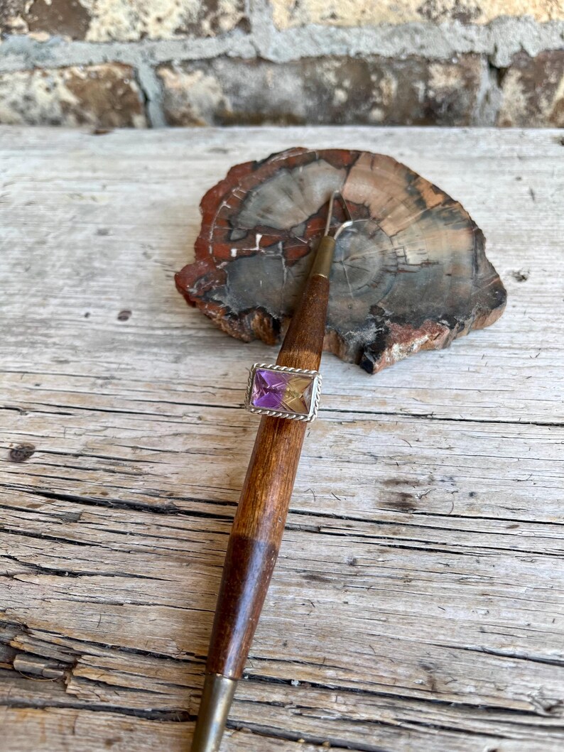 K&ouml;nnte beinhalten: Ein Silberring mit einem rechteckigen, zweifarbigen lila und gelben Edelstein. Der Ring ist auf einem Holzwerkzeug mit Metallspitze angebracht und liegt auf einem St&uuml;ck versteinertem Holz. Der Hintergrund ist eine verwitterte Holzoberfl&auml;che.