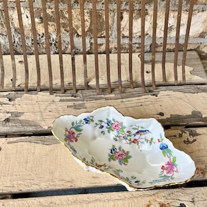 May include: A white porcelain serving dish with a scalloped edge and gold trim. The dish is decorated with floral and bird motifs in pink, blue, and green. The dish is on a weathered wooden surface.