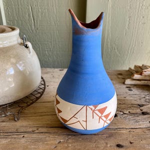 May include: A blue ceramic vase with a white band featuring geometric designs in brown. The vase has a narrow neck and a flared opening with a reddish-brown interior. The vase is on a wooden surface.
