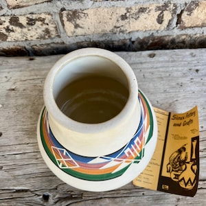 May include: Cream-coloured pottery vase with a wide opening and a decorative band featuring geometric patterns in orange, green, blue, and white. The vase sits on a wooden surface, with a brochure that reads "Sioux Pottery and Crafts".