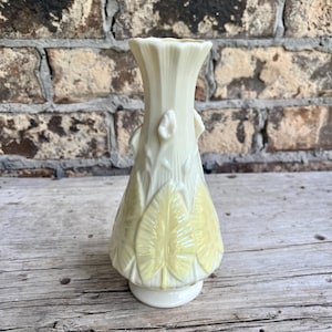 May include: A cream-colored glass vase featuring a flared, scalloped rim and a tapered neck. The body of the vase is decorated with raised, pale yellow leaf motifs and vertical ridges that extend from the base toward the neck of the vessel.
