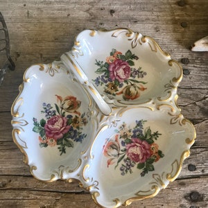 May include: A white porcelain serving dish with three sections. Each section features a floral design with pink roses, yellow tulips, and purple flowers. The dish has a gold trim and a handle.