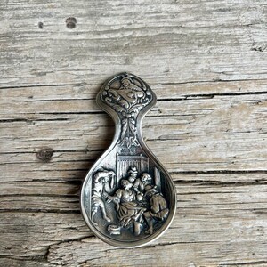 May include: A silver-colored spoon rest with a detailed relief scene of people gathered indoors. The handle features an ornate design with a cherub. The spoon rest is placed on a weathered wooden surface.