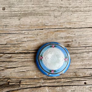 May include: A round, blue compact mirror with a floral design. The mirror has a light blue center with a white lace pattern. Pink roses and green leaves are painted around the edge of the mirror.