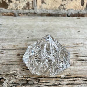 May include: A clear, faceted crystal object with a diamond-like shape. The object has a pointed top and a geometric pattern of lines and shapes. The crystal is sitting on a weathered wooden surface.