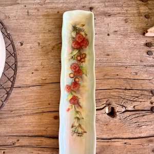 May include: A long, rectangular, white ceramic serving dish with a green glaze and a floral design. The dish features a hand-painted design of red berries and green leaves. The dish is signed "J. Gouband 1877".