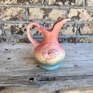 May include: A vintage pink ceramic pitcher with a handle and a floral design. The pitcher has a light blue base and a curved spout. The background is a weathered wooden surface and a brick wall.