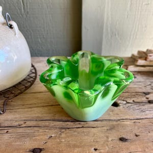 May include: A green glass ashtray with a wavy, scalloped edge. The ashtray is made of clear glass with a green tint and has a white interior.