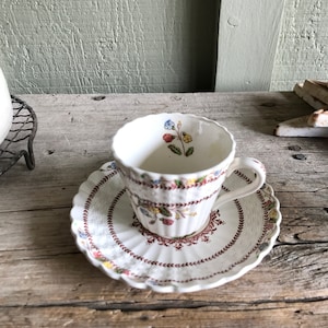 May include: A white teacup and saucer with a floral pattern in shades of red, yellow, blue, and green. The cup and saucer have a scalloped edge and a delicate floral design.