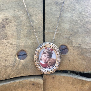 May include: A silver chain necklace with an oval-shaped locket. The locket has a gold-toned frame with a leaf design and a photo of a young girl holding a teddy bear.