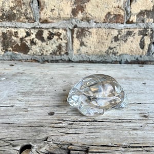 May include: A clear glass turtle figurine with detailed shell and flipper features. The figurine is displayed on a weathered wooden surface, with a brick wall in the background. The turtle is approximately 4 inches long.