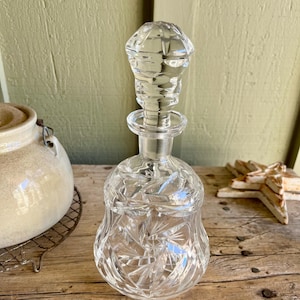 May include: A clear glass decanter with a starburst pattern and a faceted stopper. The decanter is sitting on a wooden surface.