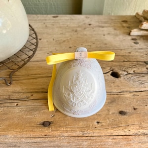 May include: A white porcelain bell ornament with a yellow ribbon and the word "LLADRO" on the top. The bell has a detailed design of a Christmas tree and the year "2003" on the front.