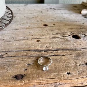 May include: A silver ring with a clear gemstone set in a prong setting. The ring is on a rustic wooden surface.